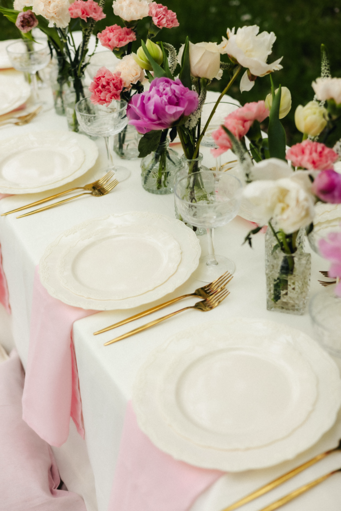 Close-up of garden party table with gold cutlery, white plates, and pink and purple peony bouquets
