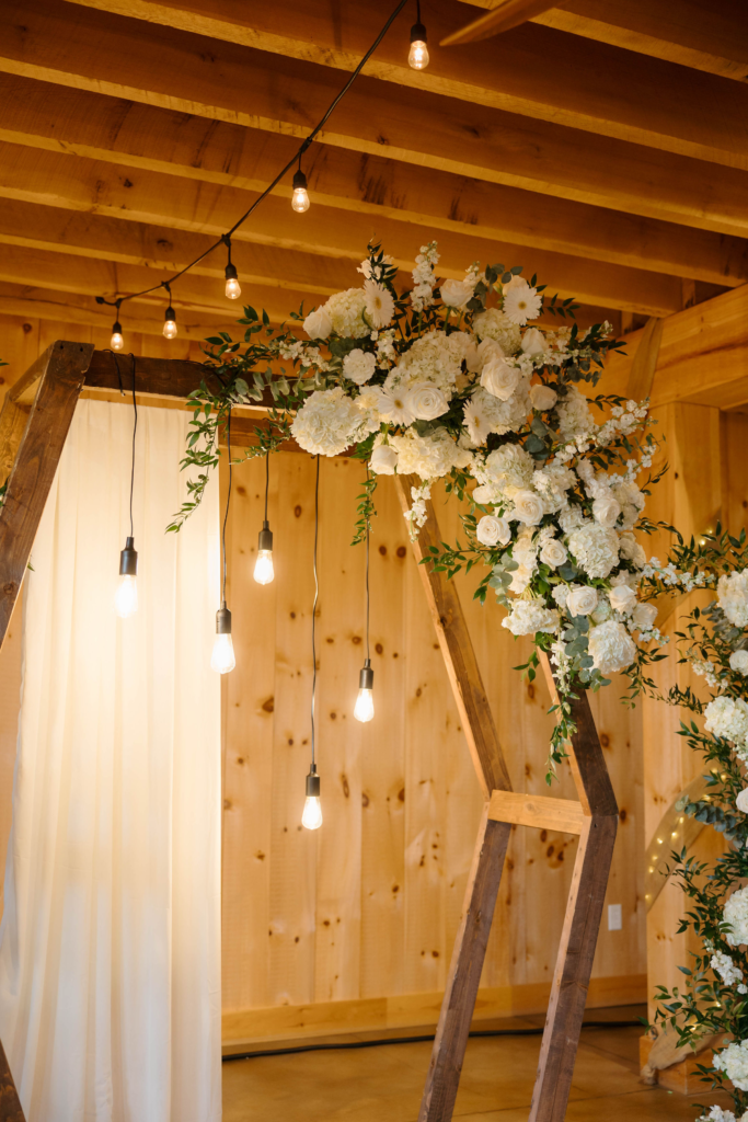 Detail shot of hexagonal wooden ceremony arch with white roses, eucalyptus, and hanging Edison bulbs