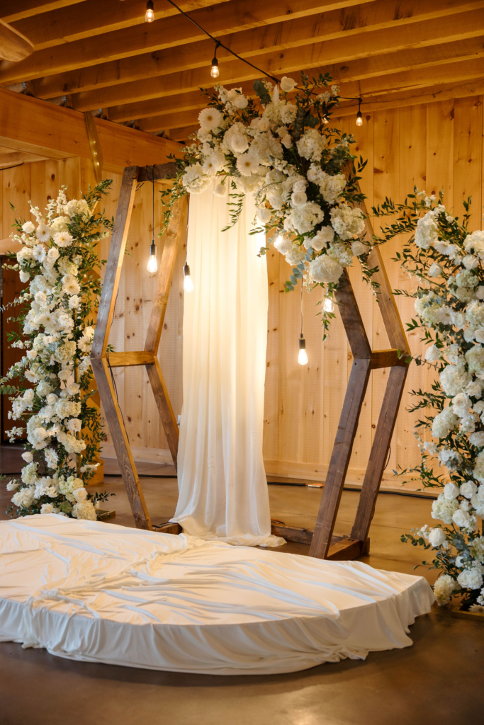 Wooden geometric wedding arch decorated with white florals and greenery in rustic barn venue