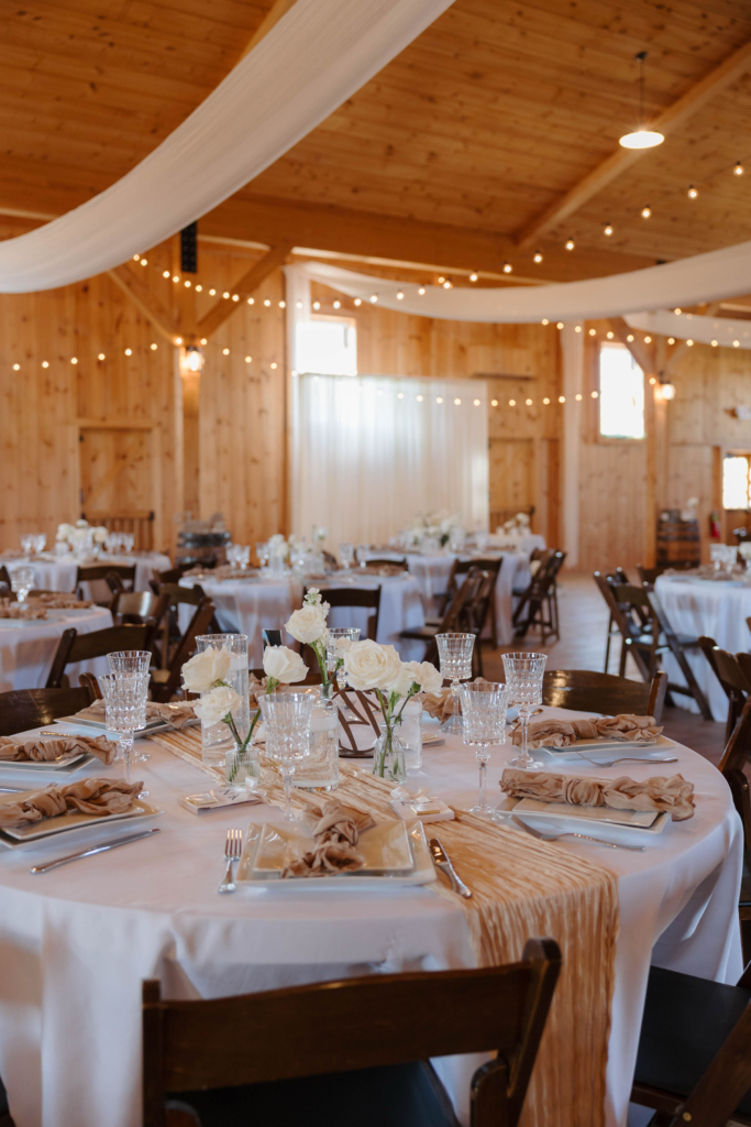 Barn wedding reception hall with string lights, white draping, round tables, and neutral linen decor