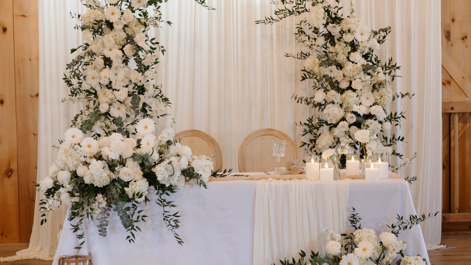 Elegant white wedding sweetheart table with rattan chairs, candlelight, and white floral backdrop