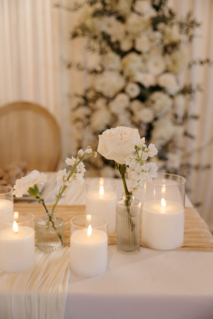 Close-up of wedding table centerpiece with white roses, candles in glass vessels, and linen runner