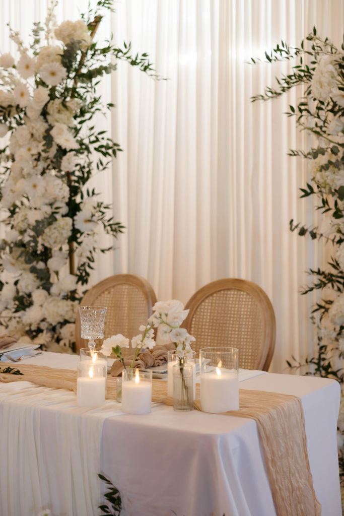 Elegant white wedding sweetheart table with rattan chairs, candlelight, and white floral backdrop